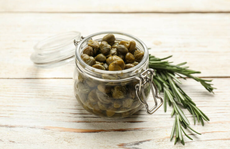 Tasty Capers In Glass Jar And Rosemary On White Wooden Table