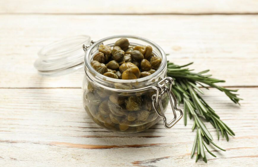 Tasty Capers In Glass Jar And Rosemary On White Wooden Table